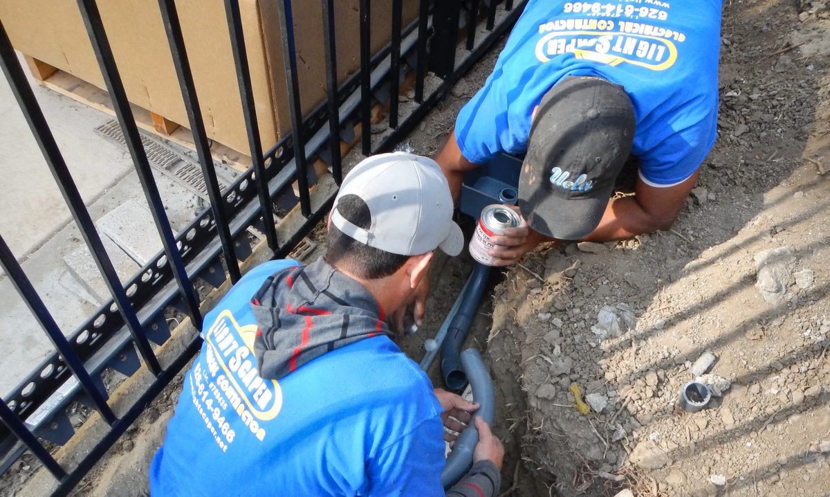 Two crew members gluing PVC conduit in a trench beside a fence